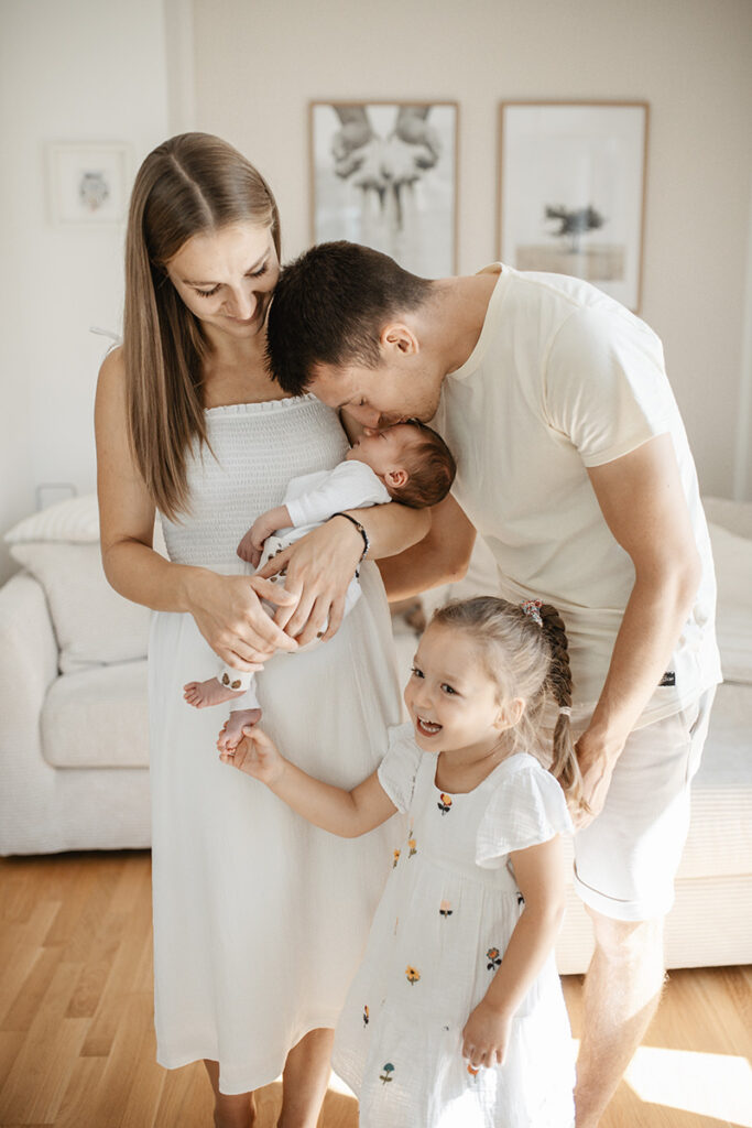 Mama hält schlafendes Baby, Papa küsst den Kopf, Geschwisterkind hält den Fuß des Neugeborenen