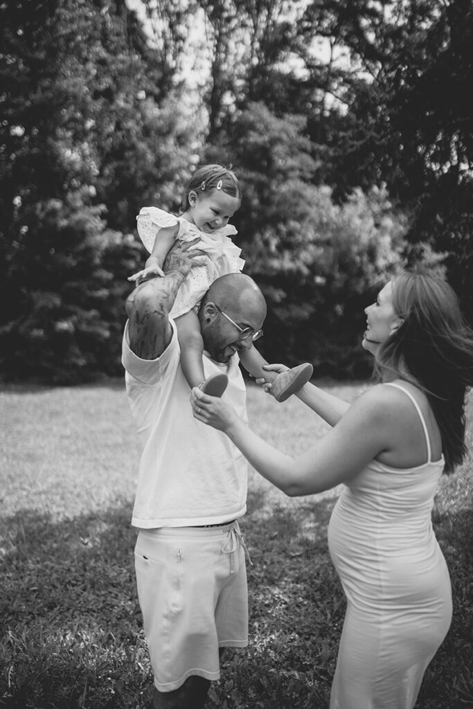 Spielerischer Moment beim Babybauchshooting im Park: Kleinkind sitzt auf Papas Schultern.