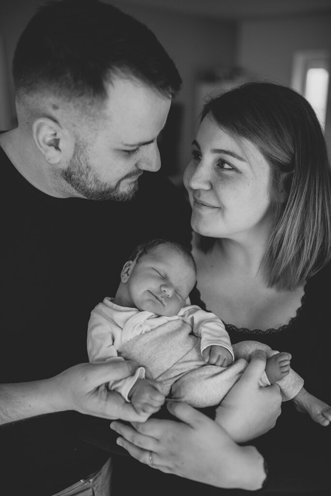 Junges Elternpaar schaut sich an, Baby schläft im Arm der Mutter – Fotoshooting für Babys