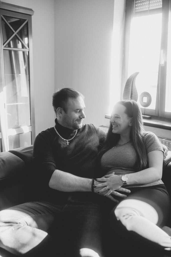 Schwarzweißaufnahme: Werdende Eltern sitzen entspannt auf der Couch bei einem Homestory-Shooting.