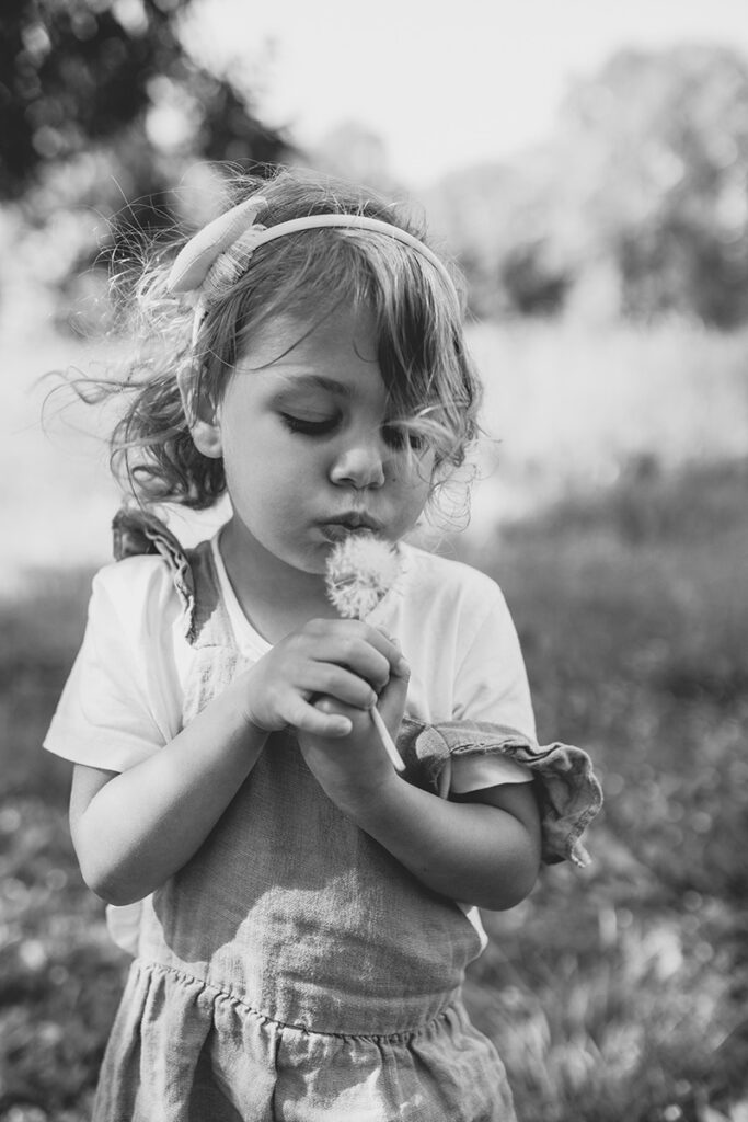 Mädchen pustet eine Pusteblume im Wind, Schwarz-Weiß-Aufnahme.