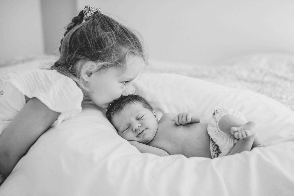 Schwarz-weiß-Foto eines schlafenden Neugeborenen, das von seiner großen Schwester auf den Kopf geküsst wird.