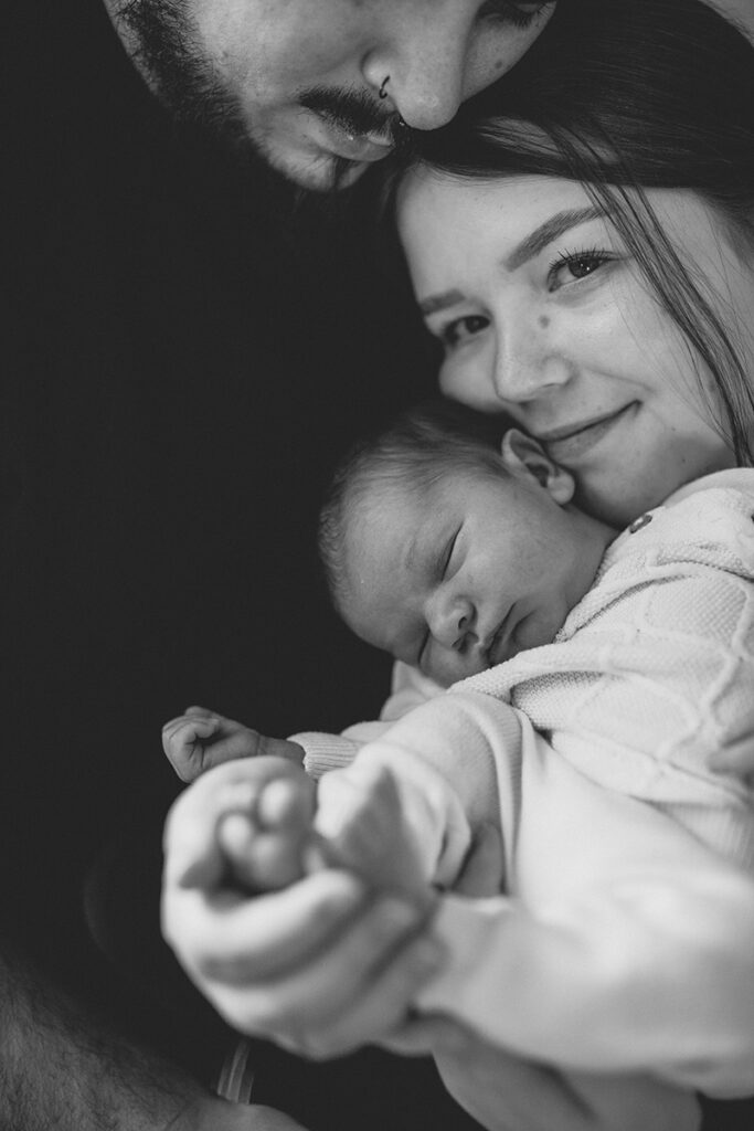 Mama hält Baby, beide schauen in die Kamera, Papa umarmt sie – newborn shooting